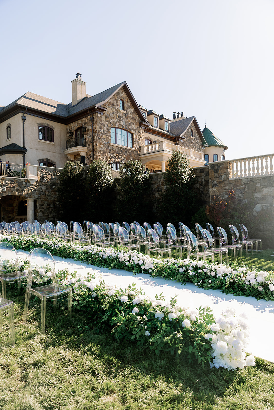 Outdoor wedding ceremony with clear ghost chairs, white floral aisle, and estate venue — Zoe Event Rentals